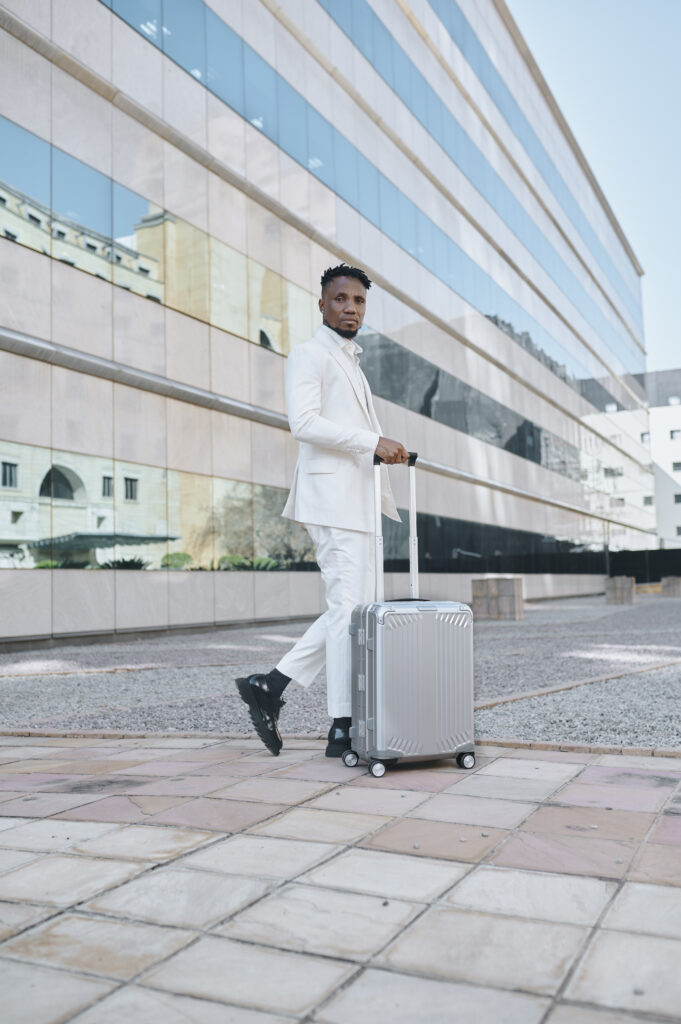 Teko Modise in white suit posing with lite-box silver suitcase